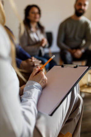 Close up shot of unrecognizable female therapist holding a clipboard for note taking during a group therapy session.